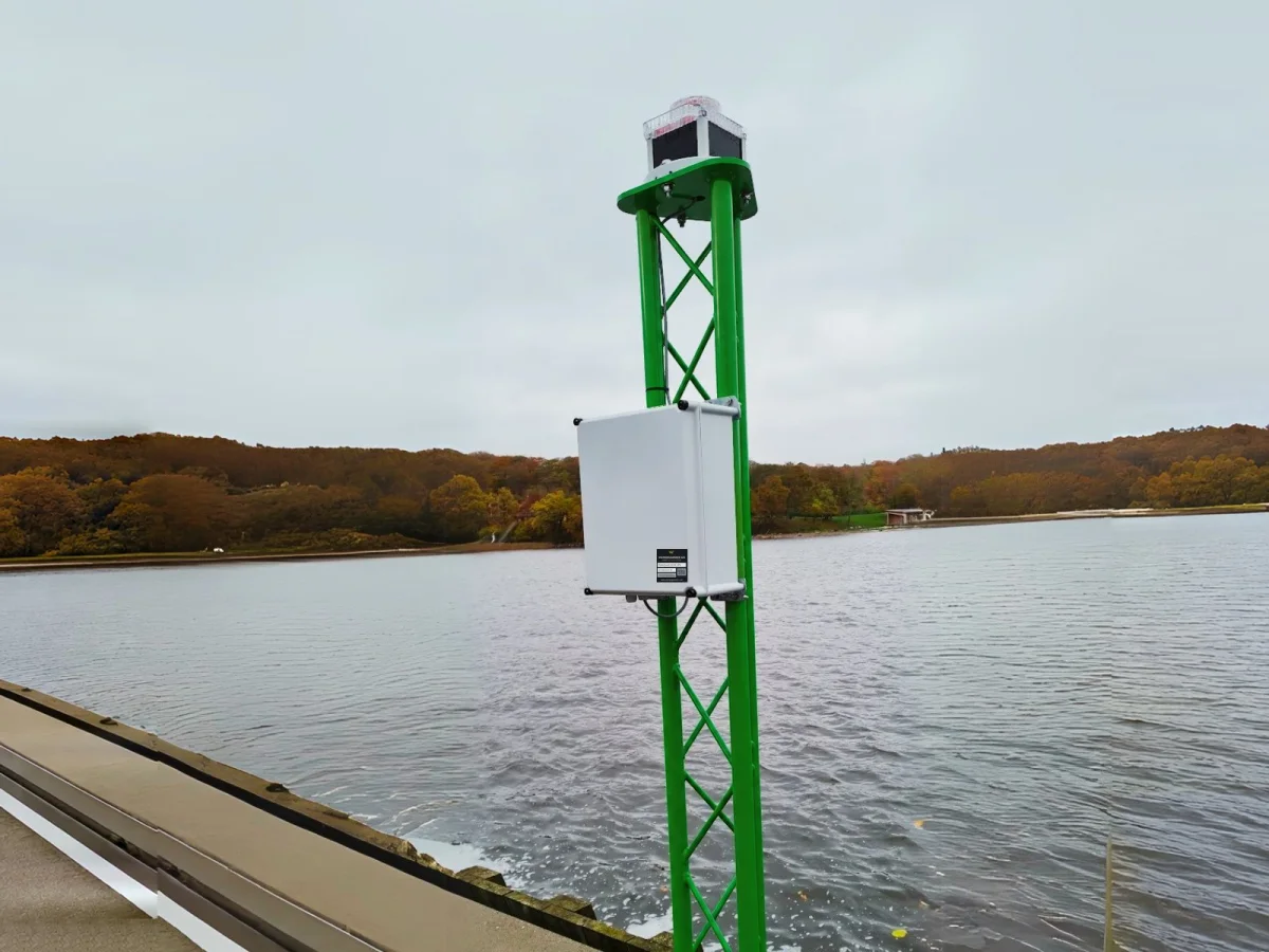 White PowerGuard enclosure mounted on a green lattice mast standing in the water, with an autumn shoreline behind.