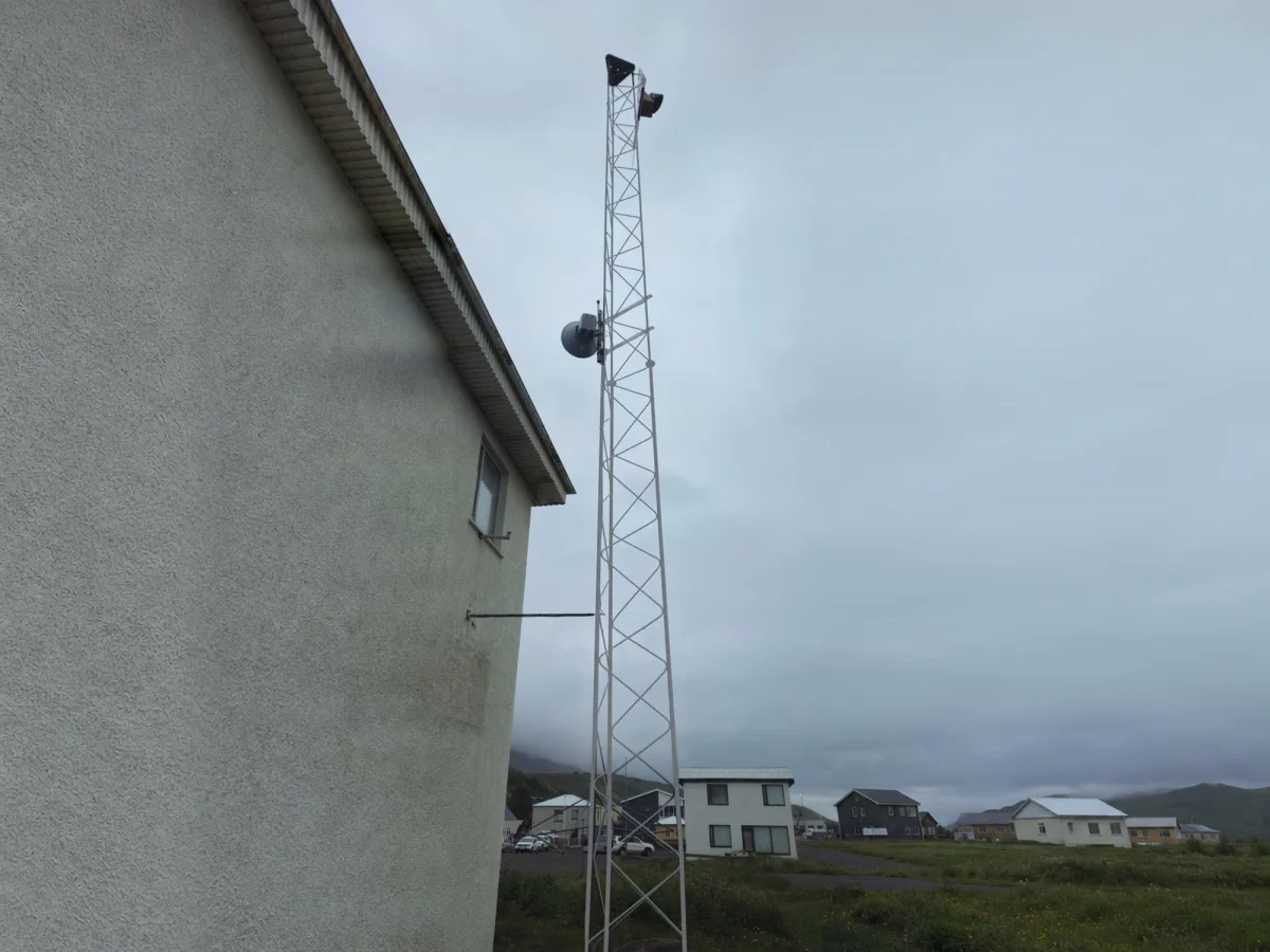 Lattice mast with a navigation light beside a harbour building, on a grey day with houses along the shore.