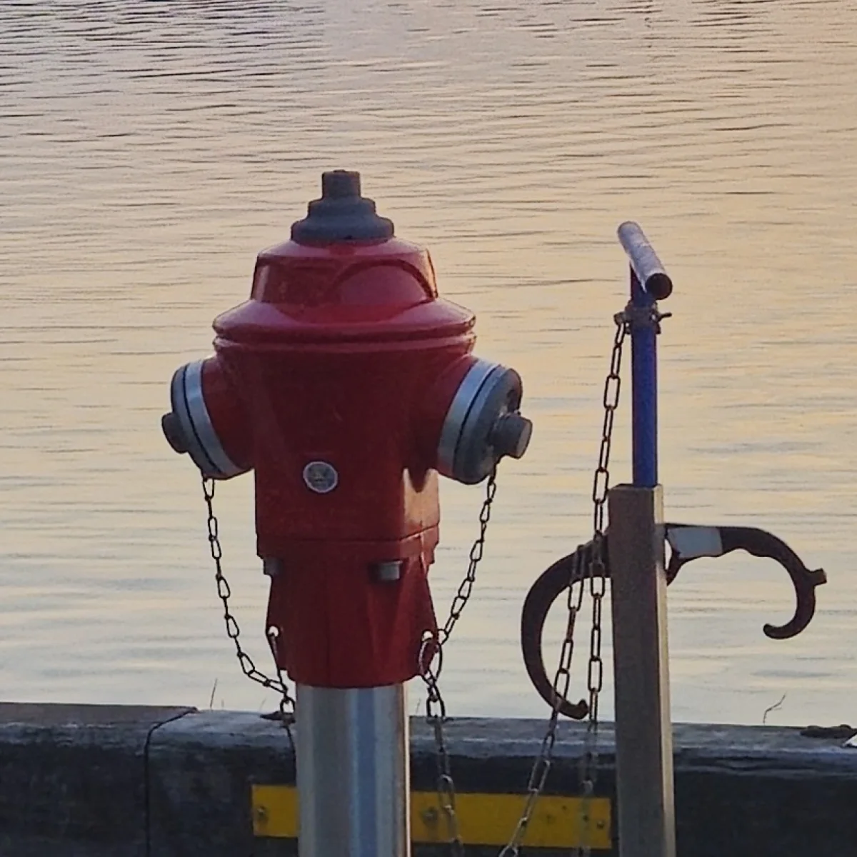Close view of the red quay hydrant with its two capped outlets and safety chains, the harbour water behind it.