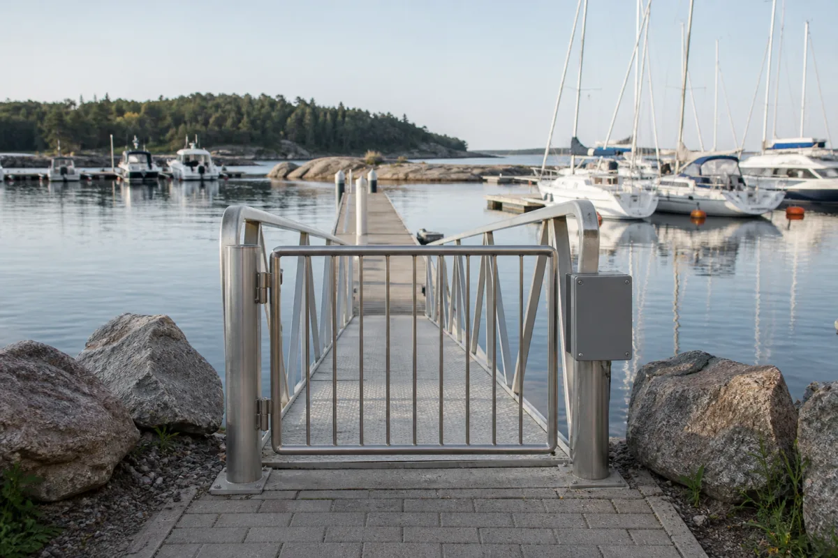 Closed stainless-steel pedestrian gate viewed from shore with a pontoon and sailboats stretching into the marina.