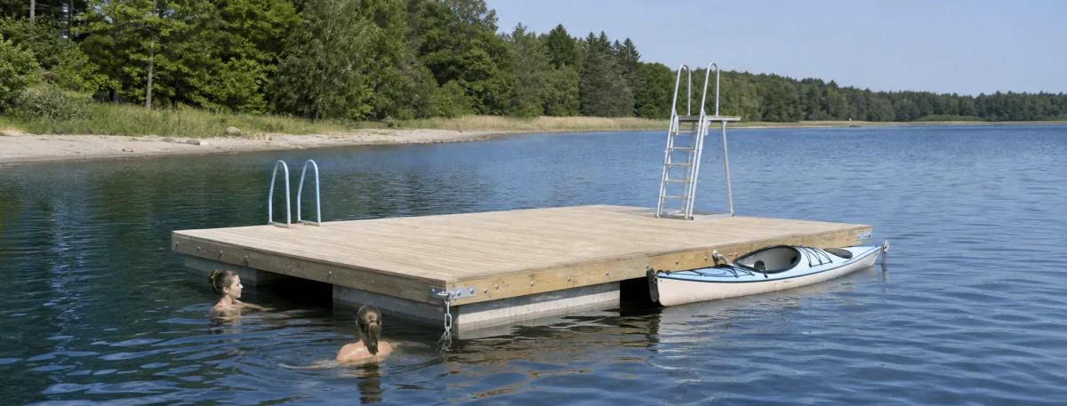 The Swim Dock is in use on a calm summer lake with swimmers and a kayak alongside.