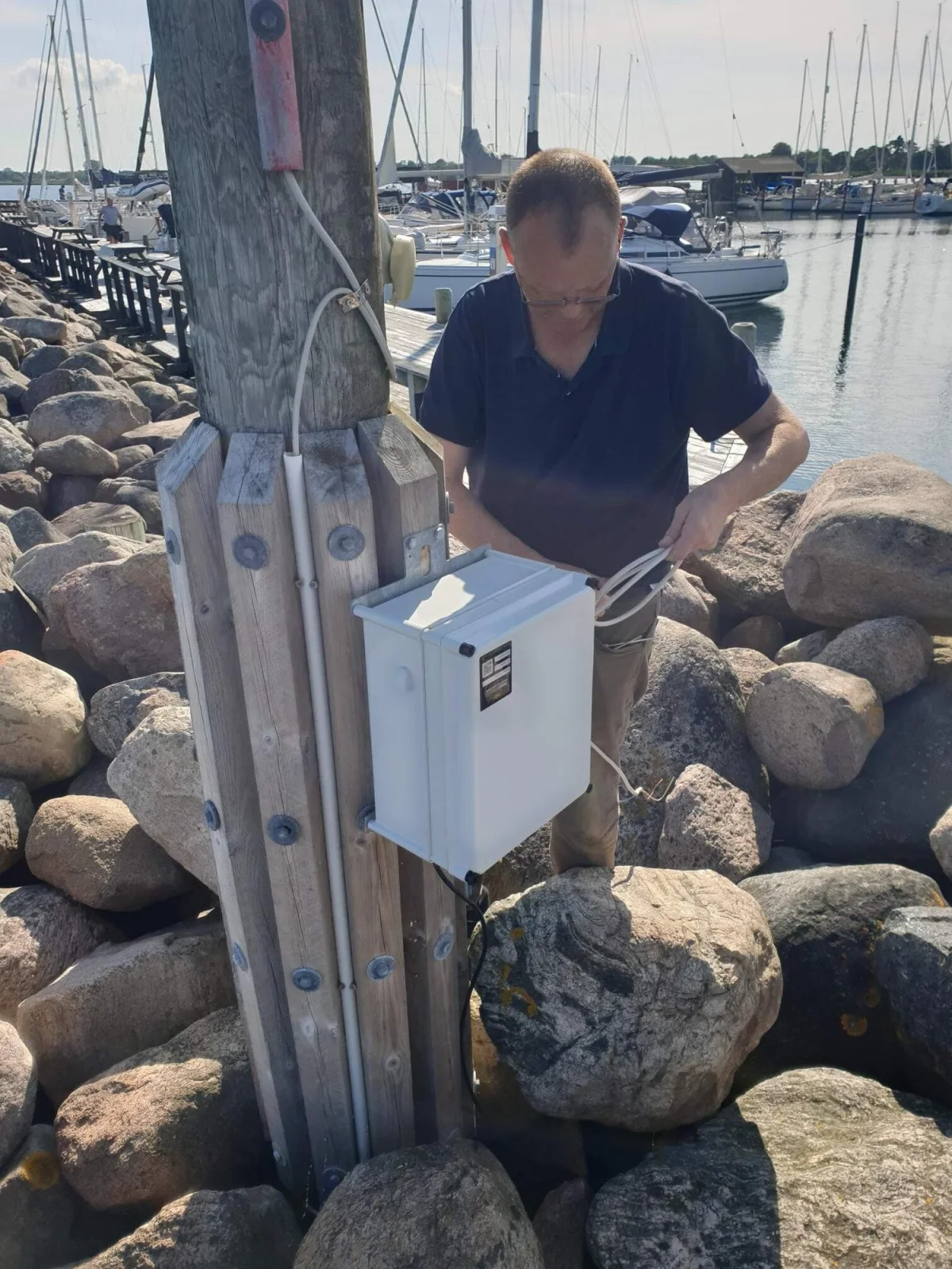 Technician checking the OceanWatcher controller box at the post, with masts in the marina behind.