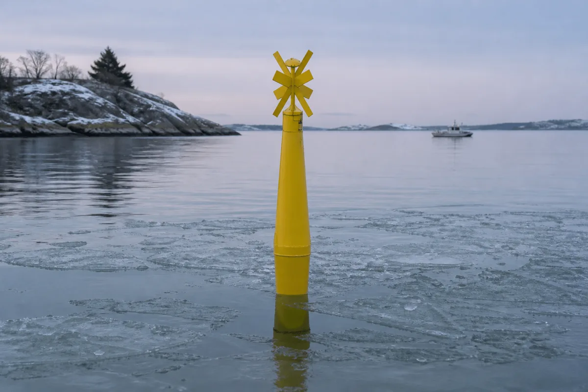 Physical Buoys yellow spar buoy standing among thin ice in a quiet Nordic fjord at blue hour.