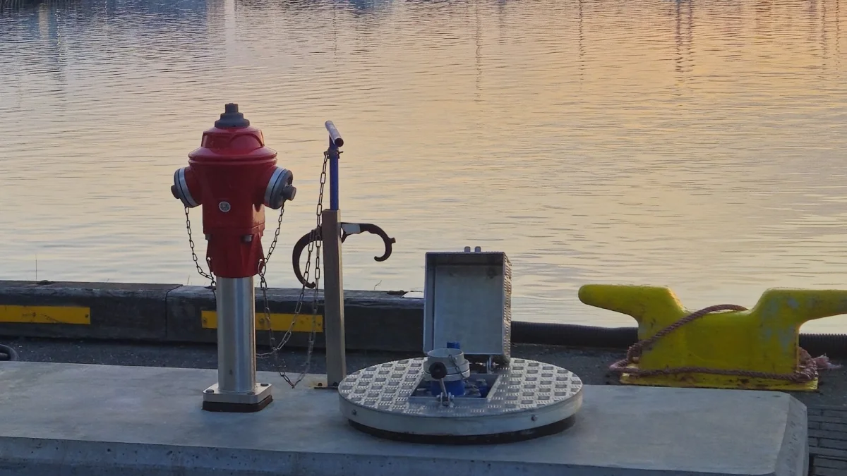 The water fittings on the quay at sunset: the red hydrant, the standpipe with its hose bracket and the open flush service point beside a mooring bollard.