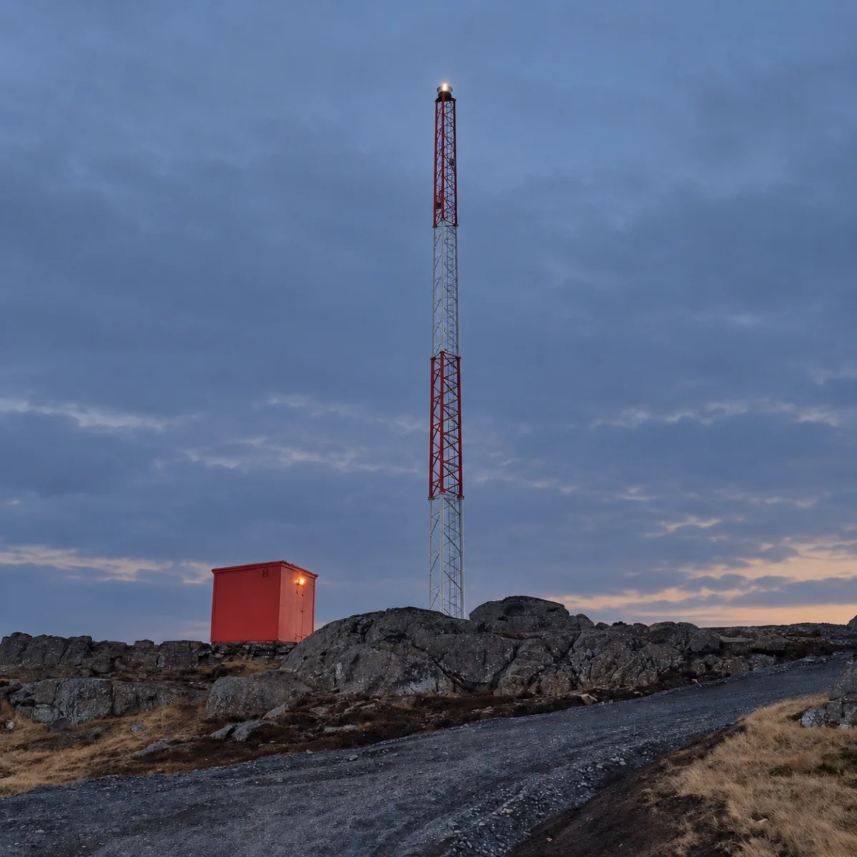 MarineGuard mast at dusk with its lantern lit, the red equipment hut beside it on the rock and the service track running up to them.