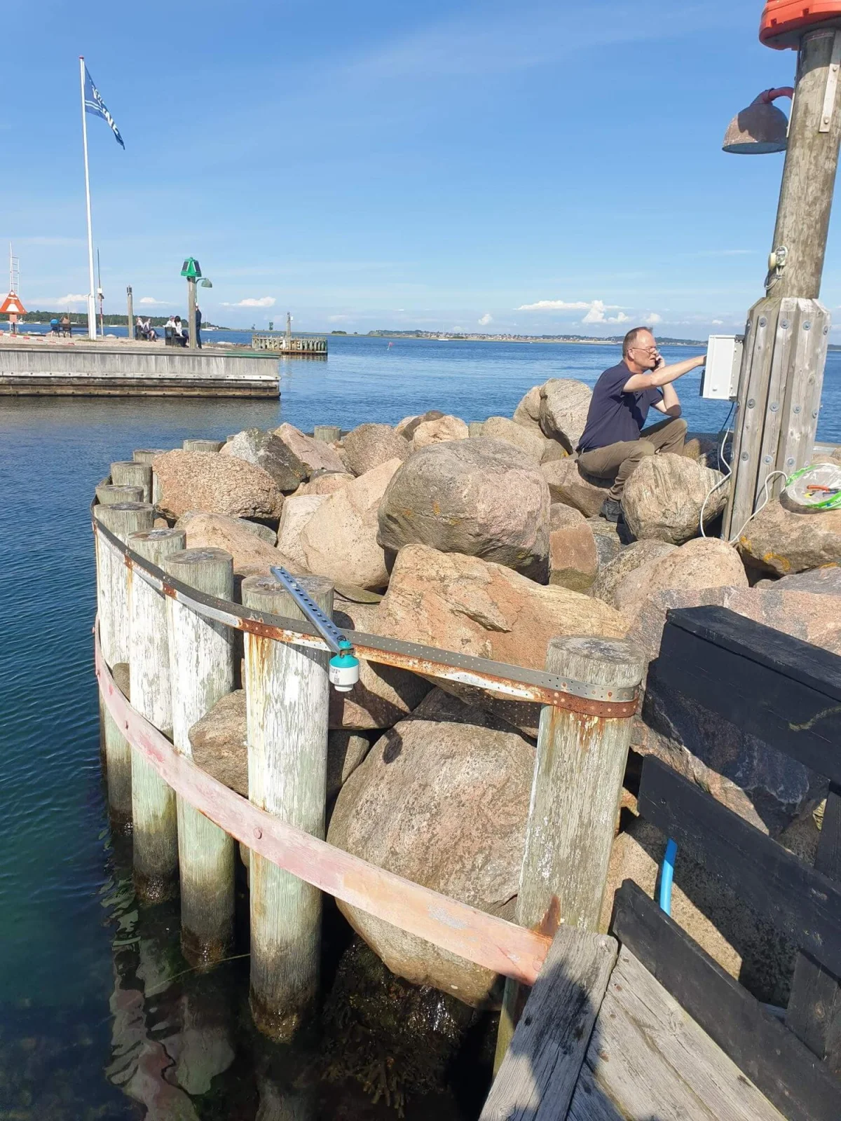 OceanWatcher arm and radar sensor on the timber breakwater, seen from the shore side over the rocks.