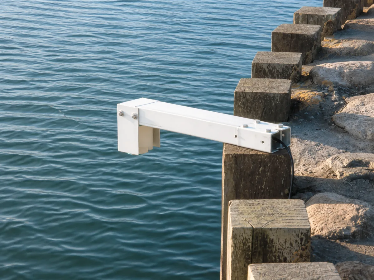 OceanWatcher radar sensor enclosed in its white aluminium arm, cantilevered over the water from timber posts at a Danish harbor edge.