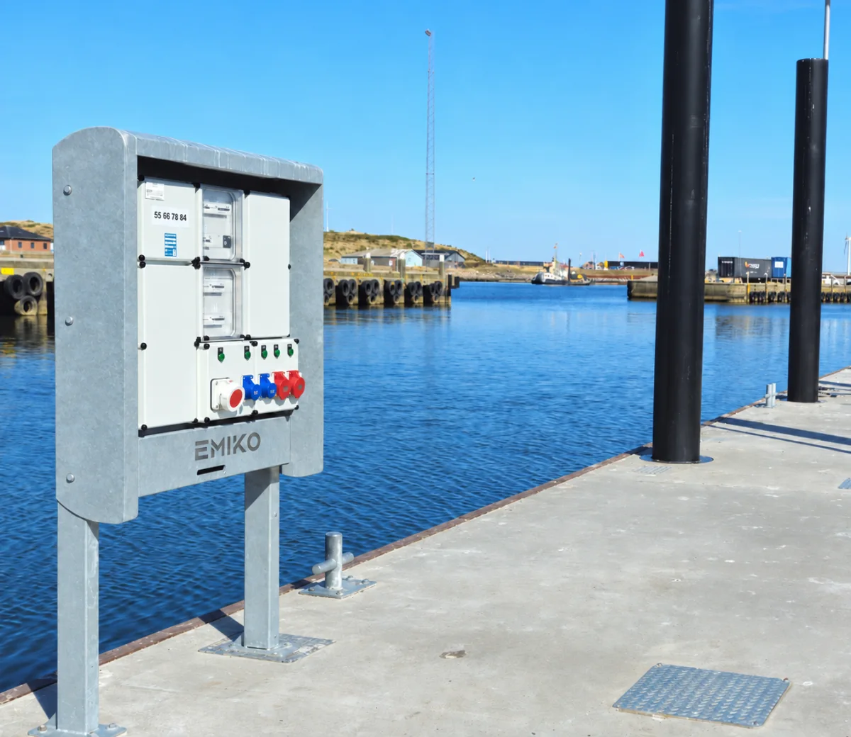 Marineium supply cabinet on a concrete quay in a Danish harbour, its galvanised frame carrying the EMIKO front with meters, breakers and red and blue CEE sockets.