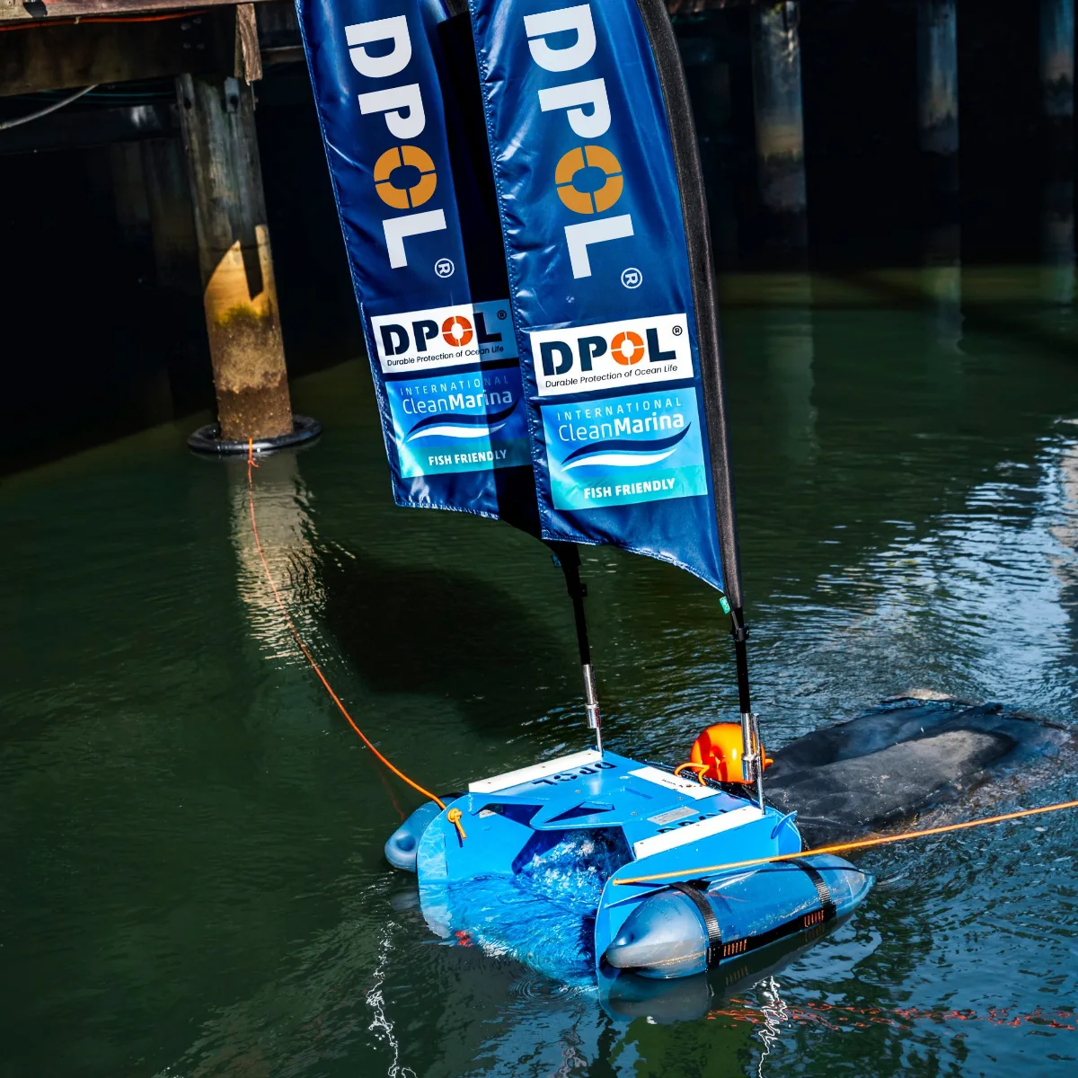 A DPOL robot collects floating waste beside a pier, flying two DPOL banner flags on the water.