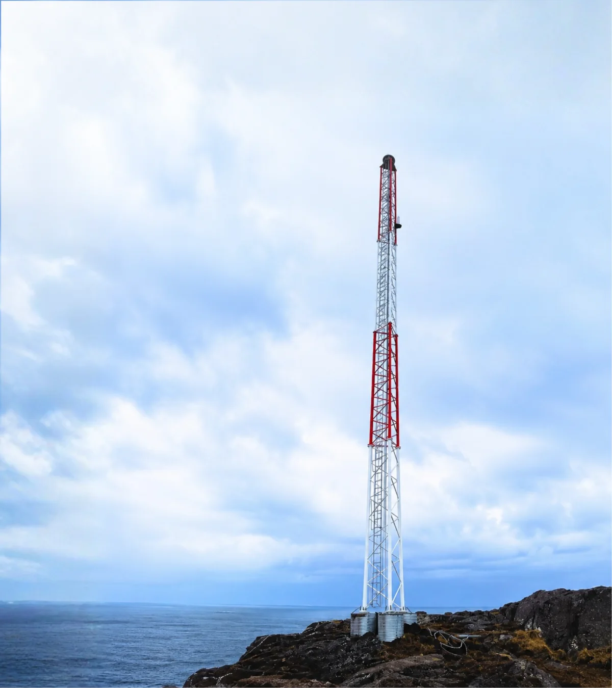 MarineGuard lattice mast standing on a rocky headland above the sea, its red and white sections carrying the lantern at the top.