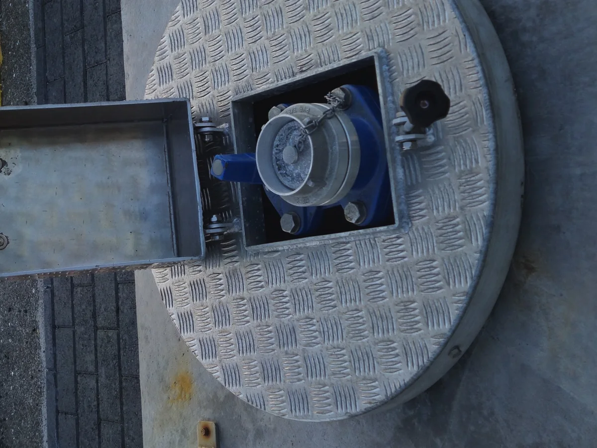 The flush service point opened: the hinged lid folded back and the blue water valve and coupling sitting in the recess under the checker plate cover.