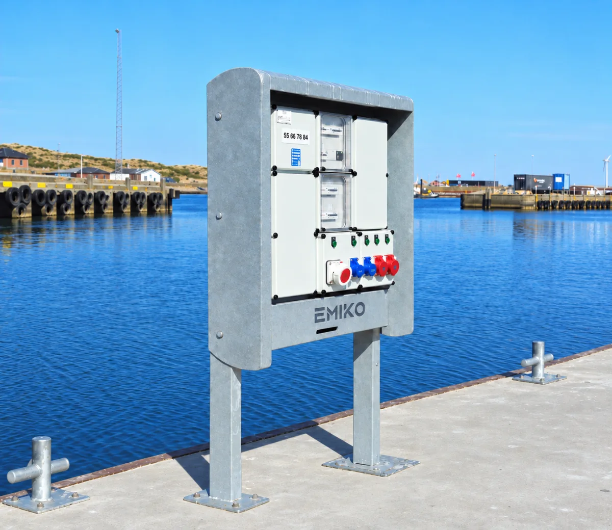Marineium cabinet centred on a concrete quay in a Danish harbour on a clear day, its EMIKO front showing the meters, the green switches and the red and blue CEE sockets.