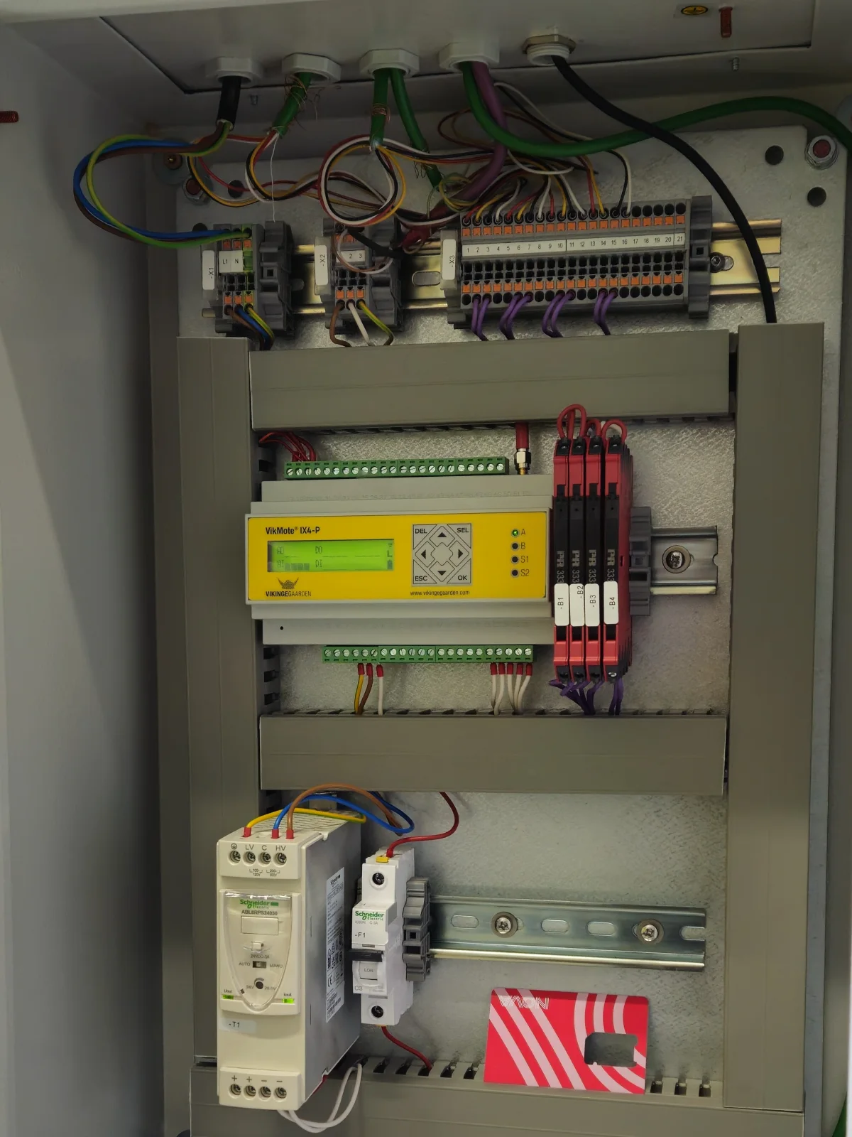 Vægskab åbnet ud for den gule VikMote-controller, forbundet til klemrækkerne ovenover og til automatsikringer og strømforsyning nedenunder, den enhed der rapporterer til Emiko-platformen.