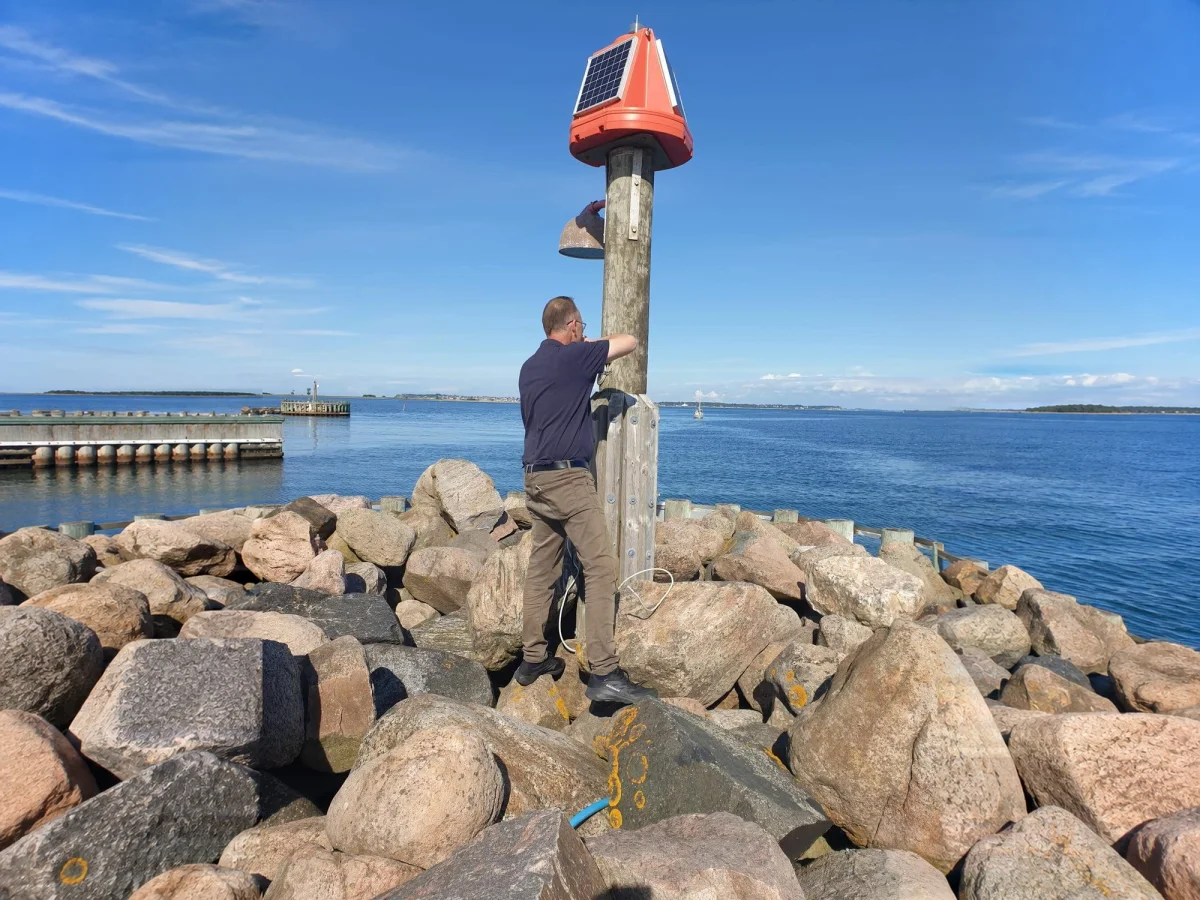 Timber post carrying a red navigation lantern and the OceanWatcher sensor, on a rocky harbour breakwater.
