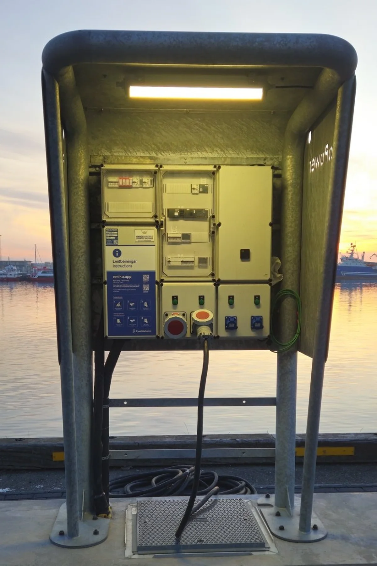 MarinePro pedestal seen from the front with its canopy lamp on and the door open on the meters, breakers and CEE sockets, the harbour at sunset behind.