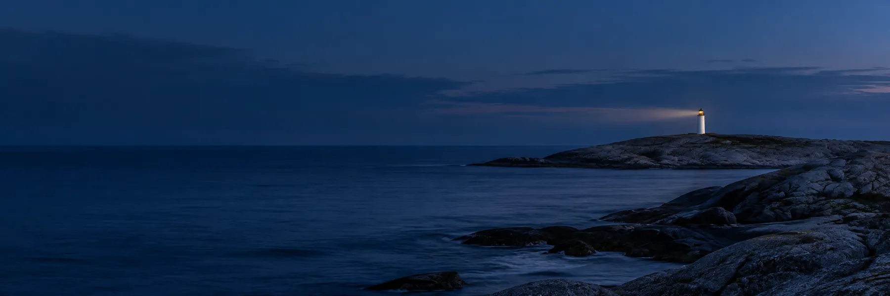 Et fjernt fyr på et granitnæs i den blå time, med lanternen tændt